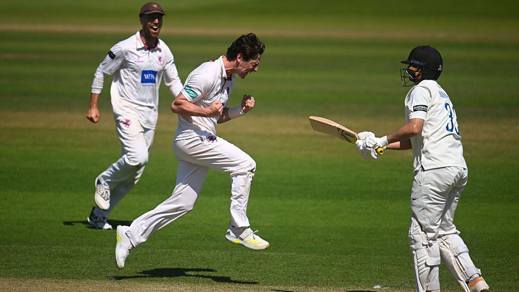 Three men in white cricket kits are on a field. The man in the middle is captured jumping into the air and celebrating. The man behind him is laughing.