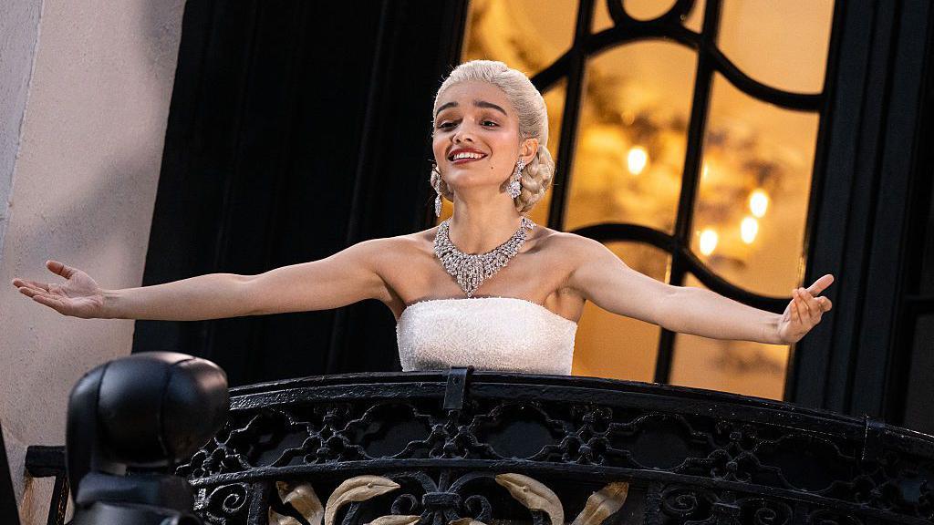 Actor Rachel Zegler performing Don't Cry For Me Argentina live on the balcony as she plays Eva Peron in Evita at the London Palladium. She has blonde hair in an elegant, low braided bun and it wearing a strapless white dress and an ornate diamond necklace. Her arms are stretched out and she is smiling while singing.