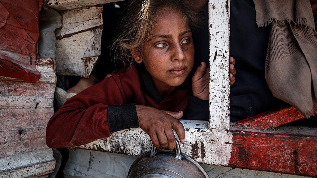 A young girl in a long-sleeved burgundy sweatshirt leans into a white and red metal barrier while holding a metal basin with her right hand as she waits to collect food at a charity kitchen in Gaza City