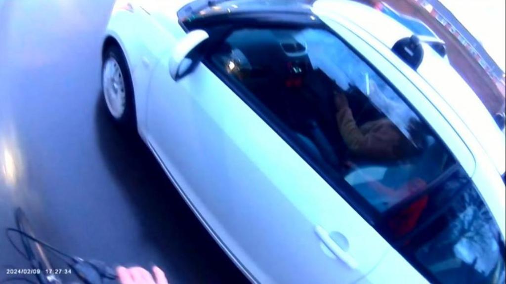 An image taken from a cyclist's helmetcam of a person driving a white car while using their mobile phone behind the wheel.