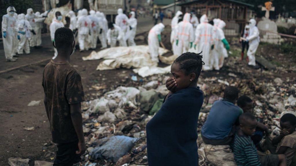 Youngsters in Goma watch as undertakers in white protective gear surround similarly white body bags