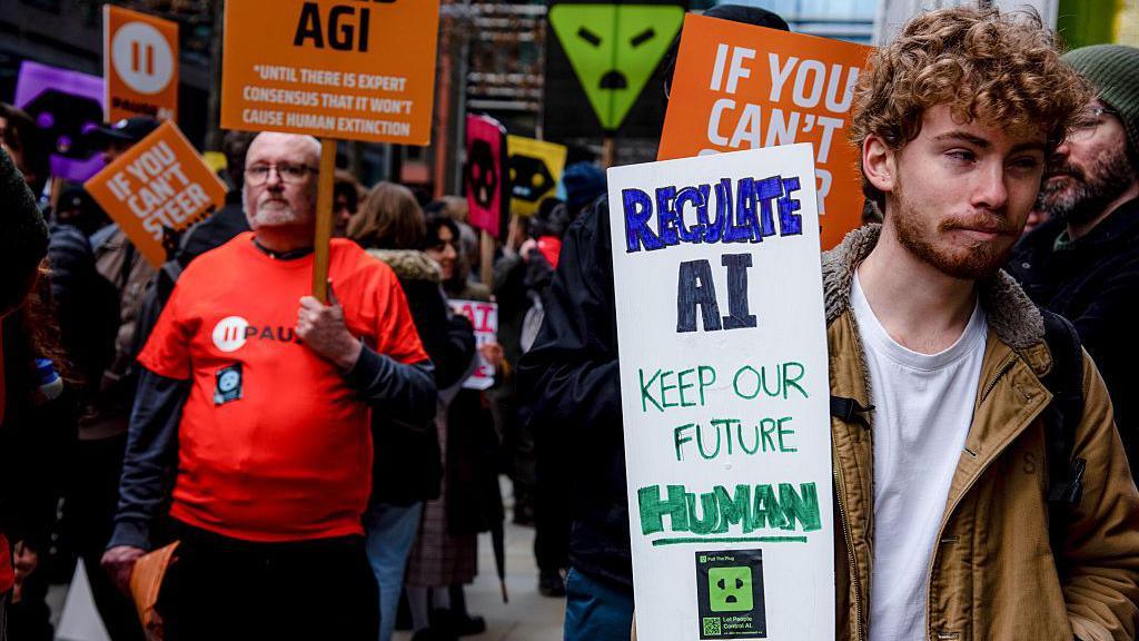 Protestwyr ar y stryd yn Llundain, rhai ohonynt gyda baneri. Tuag at flaen y llun mae baner wedi'i ysgrifennu a llaw sy'n dweud "Regulate AI, Keep our future HUMAN"