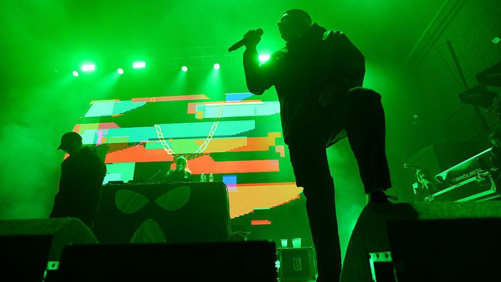 Kneecap on stage at the 02 Academy in Glasgow. Mo Chara and Moglai Bap are at the front of the stage, with DJ Provai behind on the decks. There is a colourful backdrop and the stage is bathed in green light.