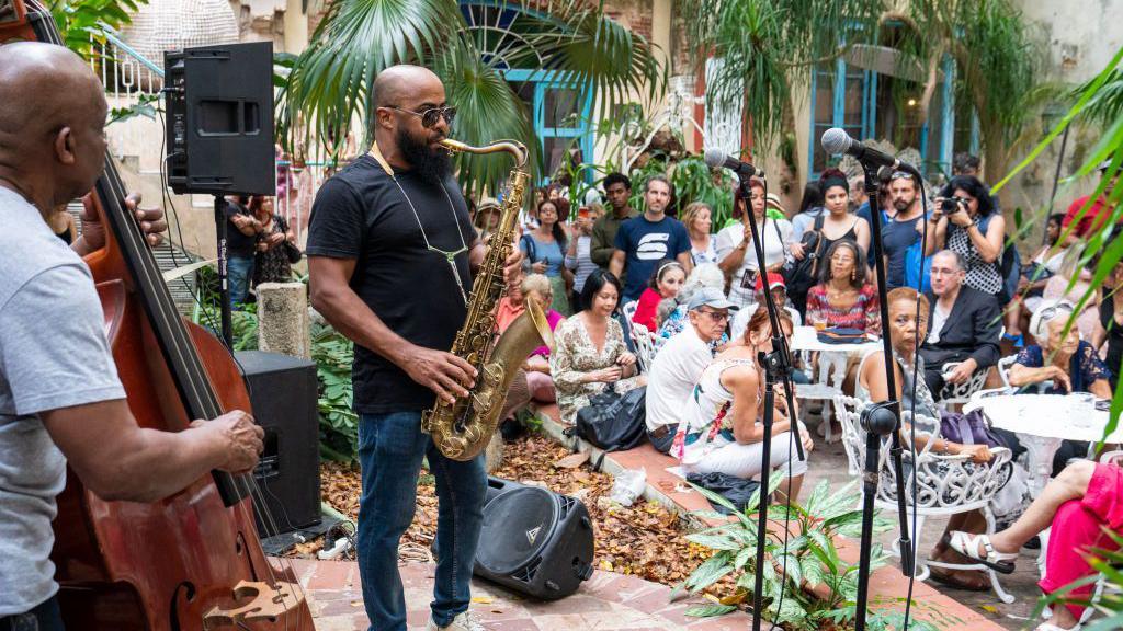 Two men are performing on a stage, one with a saxophone, another with a double bass. They are standing in a room full of green plants and audience members sitting on white garden chairs. 