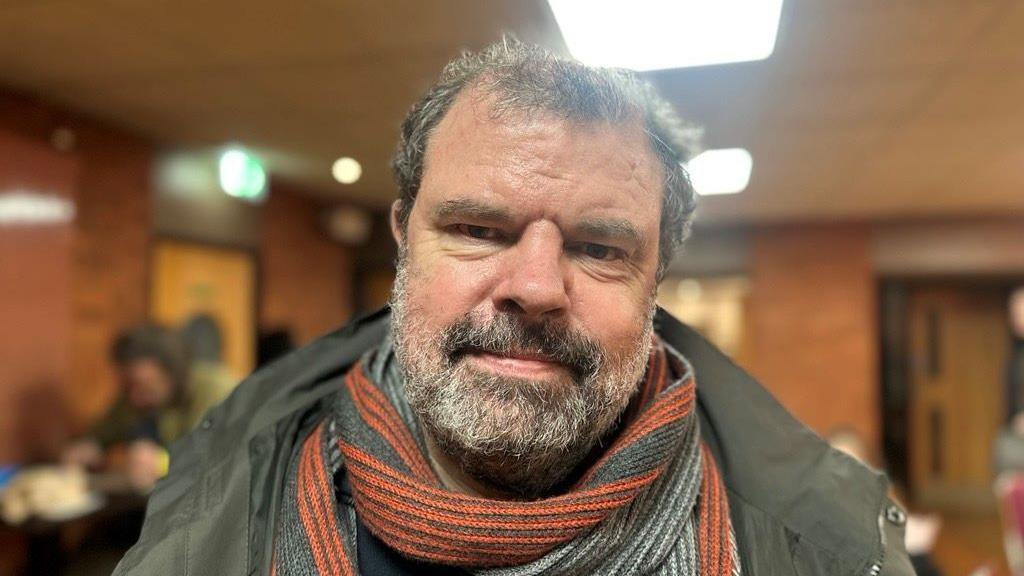 Adam lee has a black and white beard and hair wearing a ginger, grey and white scarf.
