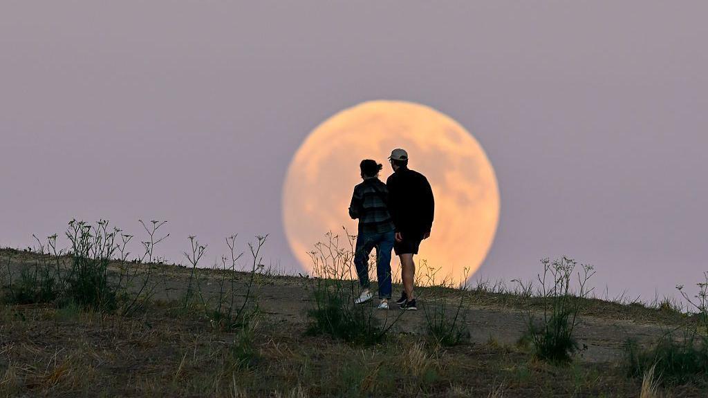 A woman and man walk on a dirt trail on a hillside in front of a full moon in a purple sky