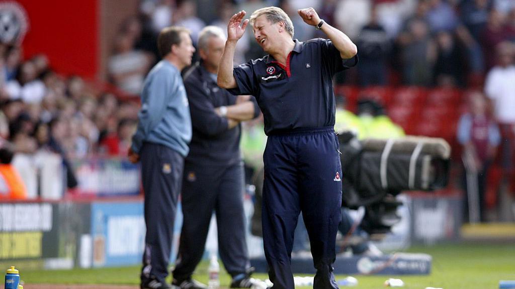 Neil Warnock, a middle-aged man with grey hair and wearing a navy blue tracksuit, laments a decision on the touchline.