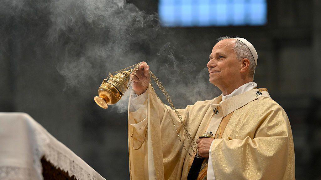 Pope Leo XIV, wearing long robes and a white skullcap, waves a gold thurible in the air, which emits smoke from incense.