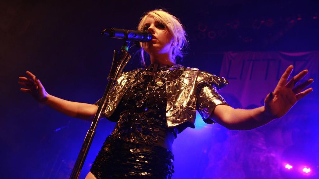 Victoria Mason performing on stage during a tour, wearing a black and gold intricate leotard with a gold cropped jacket. She has bleach blonde hair pulled back and large theatrical feather eyelashes on. She is singing into a microphone with her arms out beside her. The stage is dark and smoky with a pink and purple hue.