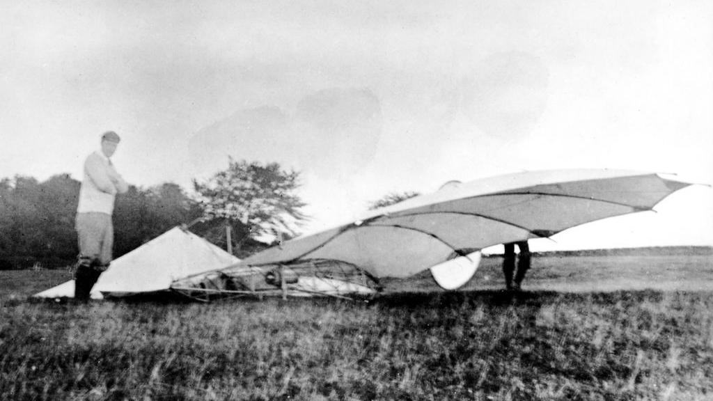 A black and white image of Percy Pilcher standing in a field next to a crashed glider.