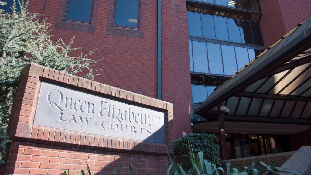 A close-up photo of a redbrick building on a sunny day. A sign at the building's entrance says "Queen Elizabeth II Law Courts." There is a hedge growing behind the sign.