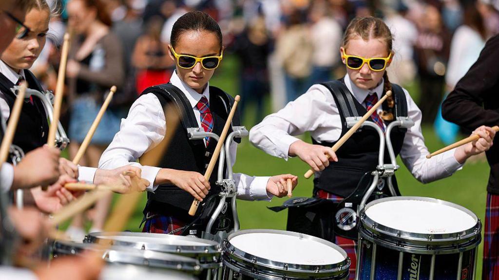 Close-up of a group of teenage girls who are marching drummers performing outdoors. They are wearing traditional outfits and their drumsticks are raised, about to come down on the drums. Spectators are blurred in the background