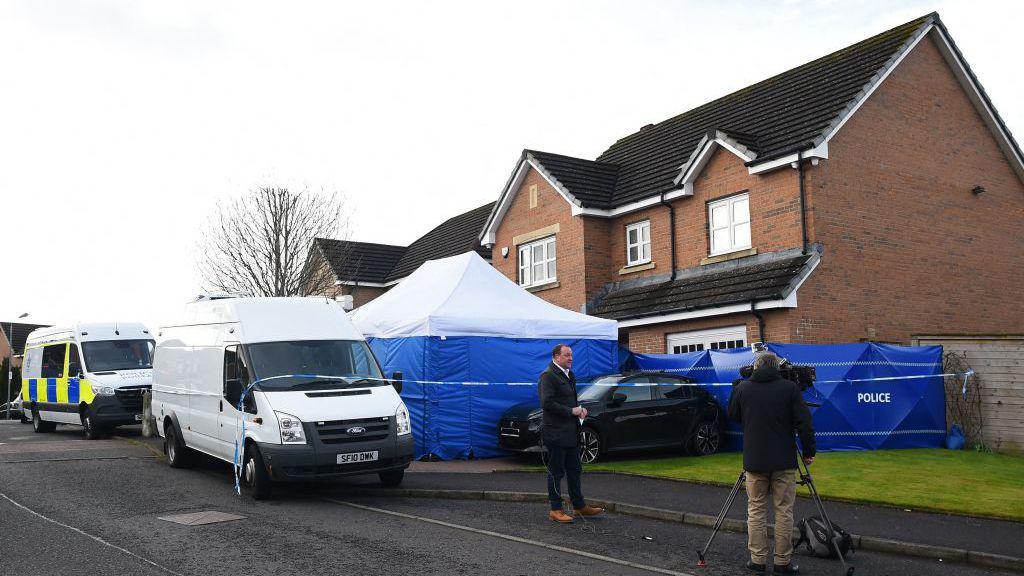 A house with a blue forensic tent in front of it. The tent has a white roof. The house is red brick and has a dark-coloured roof with white guttering around it. In front of the tent is a white van. Behind the van is another van, but with police marking on it. In the foreground, a journalist wearing dark clothing and brown shoes stands in front of a camera with a cameraman wearing a dark jacket and light trousers.