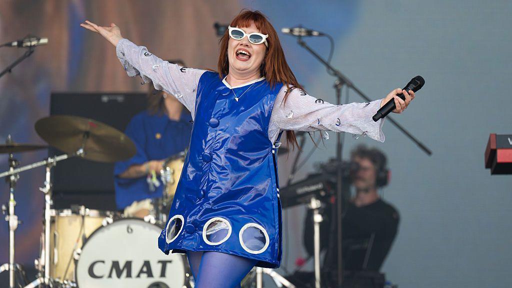 Woman on stage, in front of a set of drums, carrying a microphone and wearing white-rimmed sunglasses. She is wearing a blue plastic-looking mini-dress and blue tights. She has her arms out-stretched and is smiling widely at the crowd.