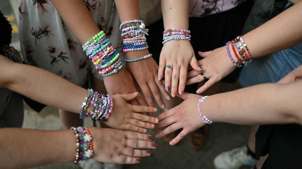 7 outstretched arms featuring Taylor Swift bracelets with the names of songs.