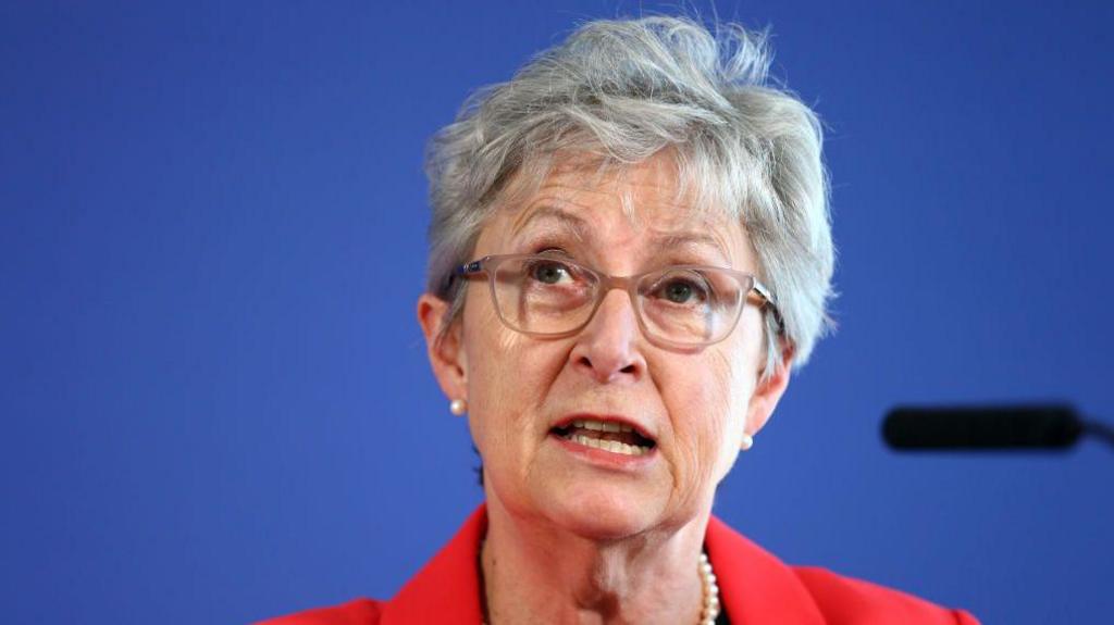 Gisela Stuart giving a speech in a red suit in front of a blue background