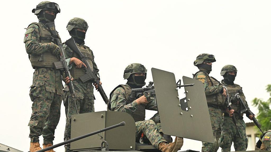Five Ecuadorean soldiers stand atop an armoured vehicle at a command post in Guayas province. They are wearing camouflage uniforms, helmets and their faces are partly covered by black balaclavas. They are all heavily armed.