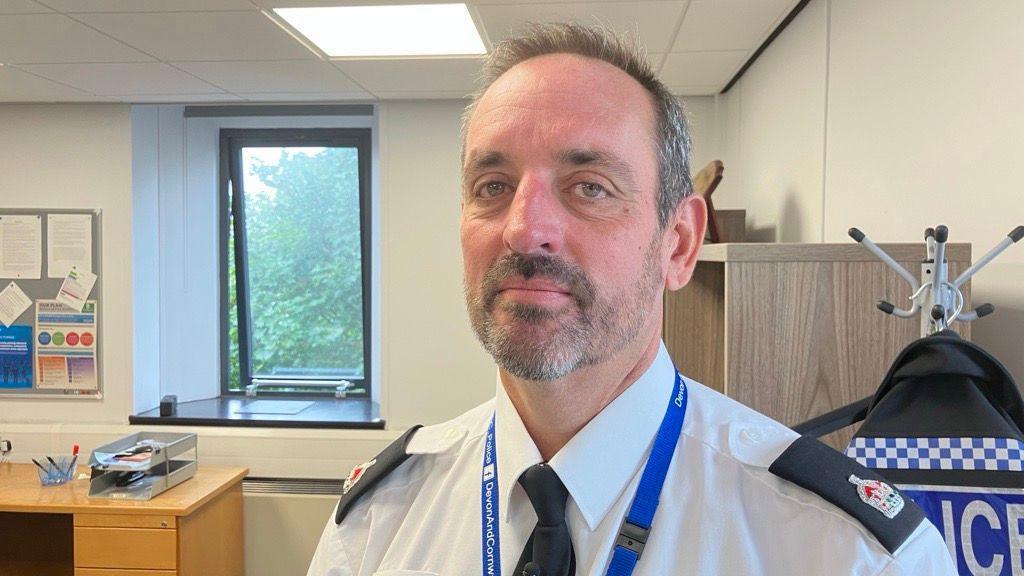 A picture of a man smiling at a camera. He is wearing a white shirt with a tie and a blue lanyard around his neck. He is standing in an office room and is in front of a coat with the word Police on it.
