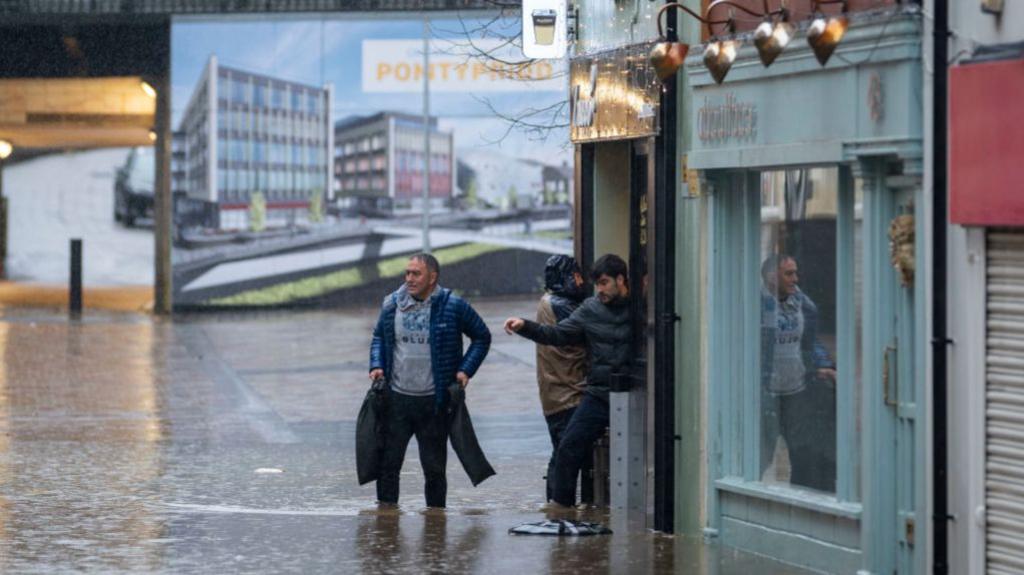 Flooding on Mill Street on November 24, 2024 in Pontypridd, Wales - a group of men stand outside some shops, with water coming up past their shins.