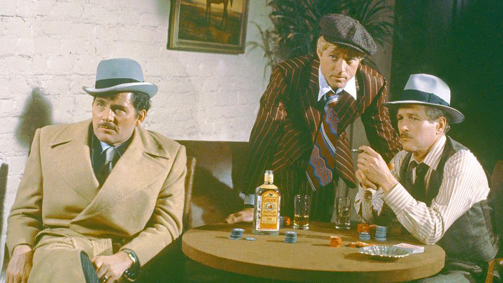 Shaw is wearing a camel coat over a light grey suit, tie and shirt and a light grey Homburg hat. He is sitting at a table with an open bottle of gin, drinks, cards and gambling chips. Robert Redford, in a brown chalk-stripe suit, blue, brown and white striped tie, flat cap and blue shirt, is leaning over the table. Paul Newman is in a grey trilby, grey waistcoat, white and brown striped shirt and is smoking a cigar while sitting at the table. 