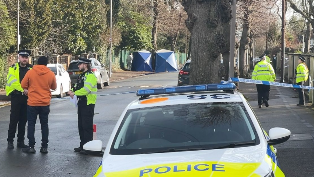 Addington Road, a tree-lined street in Reading. In the foreground is a police car. Two police officers stand near it talking to a member of the public. In the background are two blue police forensics tents.