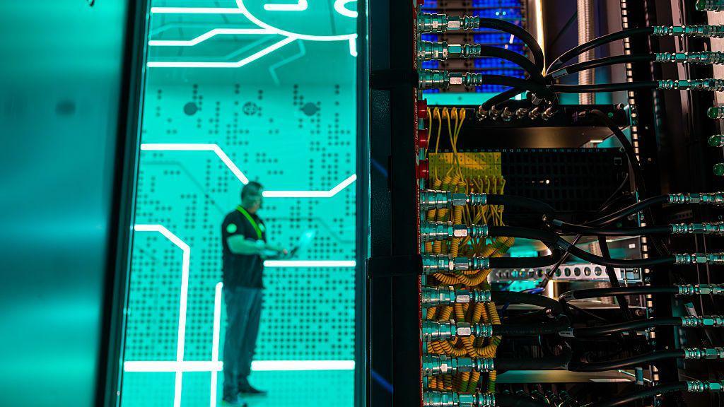 A man standing against a bright green background in a data centre - in the foreground is the back of a large computer with lots of yellow and black wires.