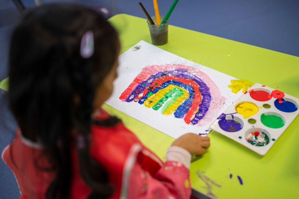 A primary school-aged girl sits at a yellow table with her painting of a rainbow. She is wearing a red apron. 