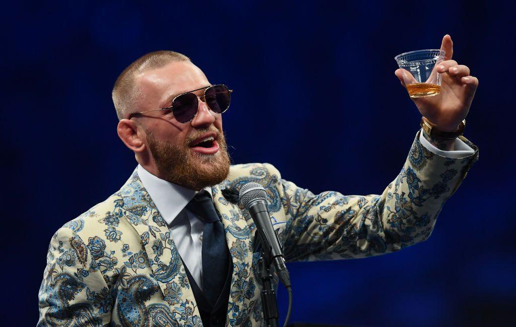 Conor McGregor in a patterned blazer, smiling, wearing sunglasses and holding up a shot of whiskey