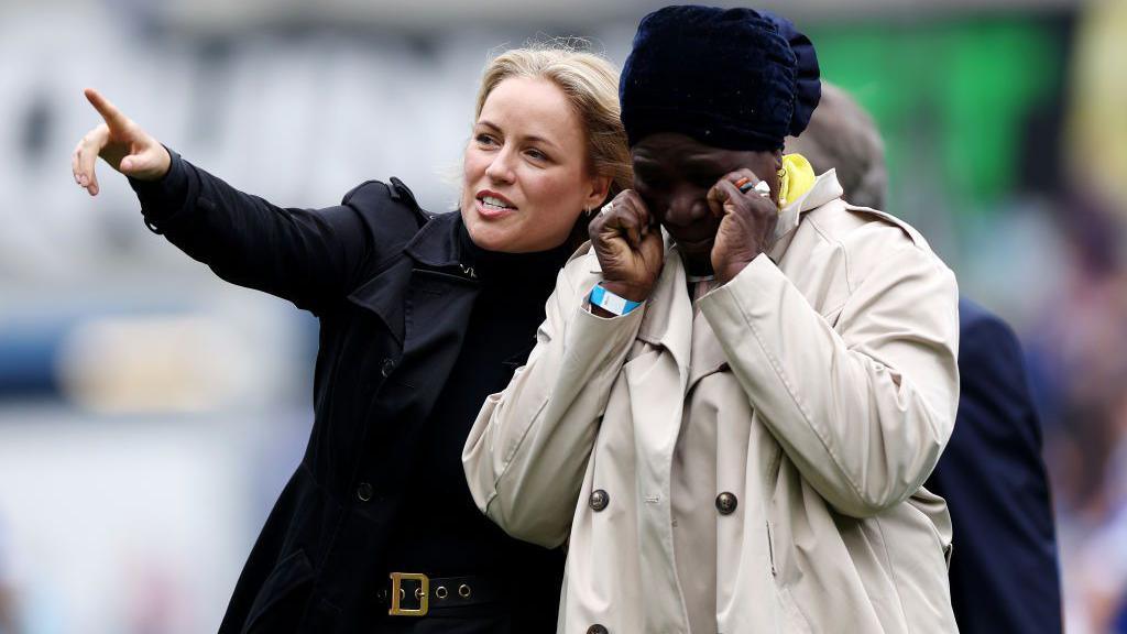 Chloe Bamba points to the crowd with her arm around Bamba's mother who has her hands to her face.