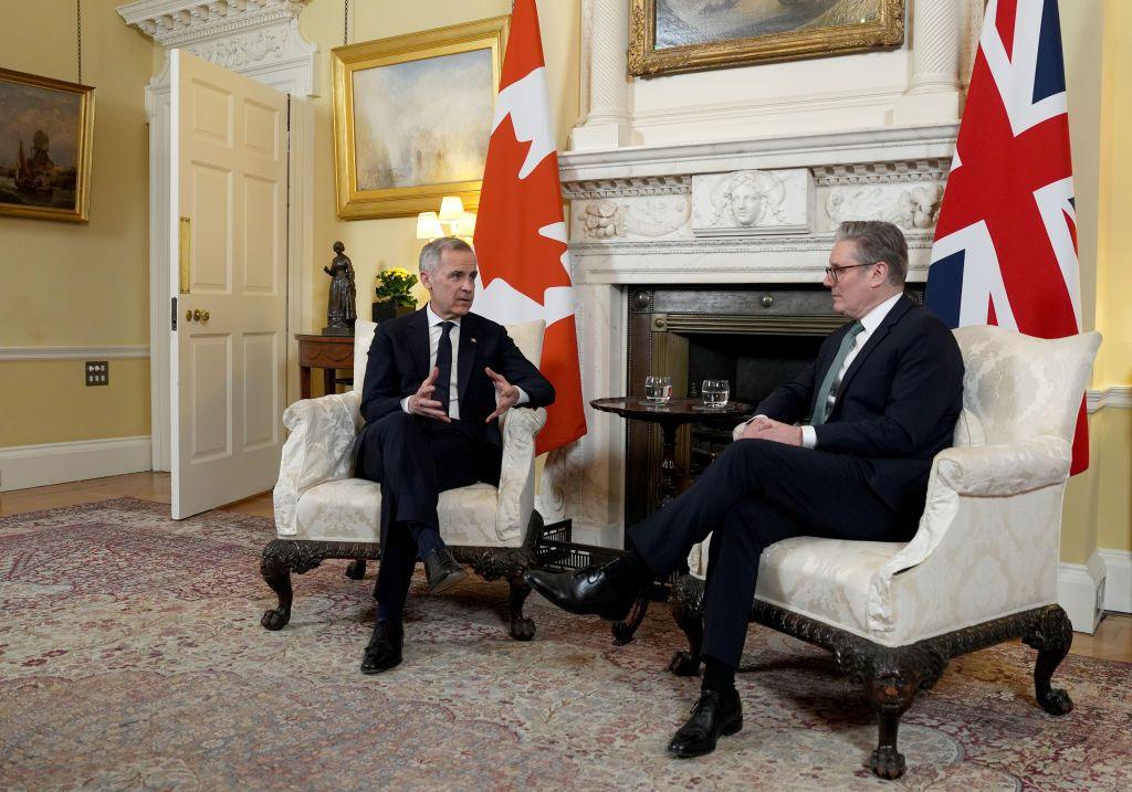 Mark Carney and Sir Keir Starmer wearing suits and sat down on white arm chairs in front of the flags of Canada and the UK.