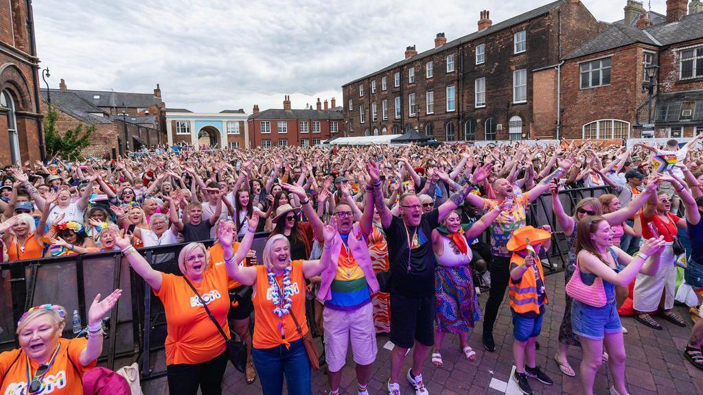 A crowd of people stood in a city centre waving their hands in the air. The sky is bright but cloudy. 