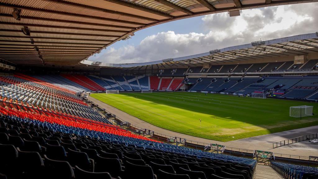 An interior shot of Hampden Stadium. The bright green grass pitch is encircled by blue and red seats. The stadium opens out at the top in an oval shape.