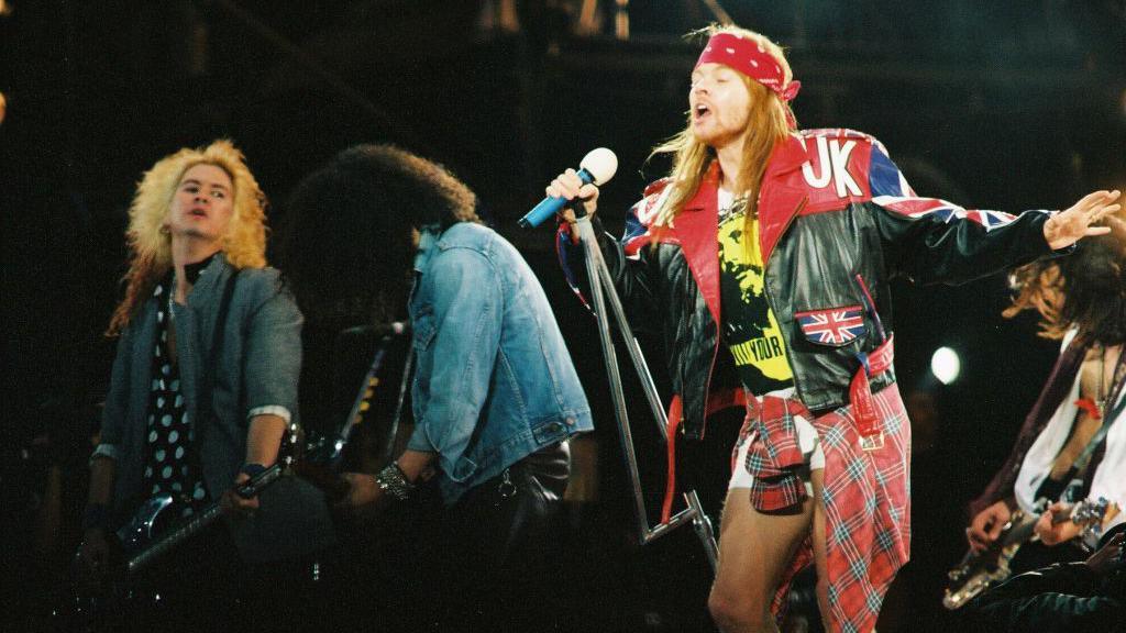 Axl Rose singing onstage wearing a bandana and a leather jacket. Two guitarists and a bassist are visible in the background. 