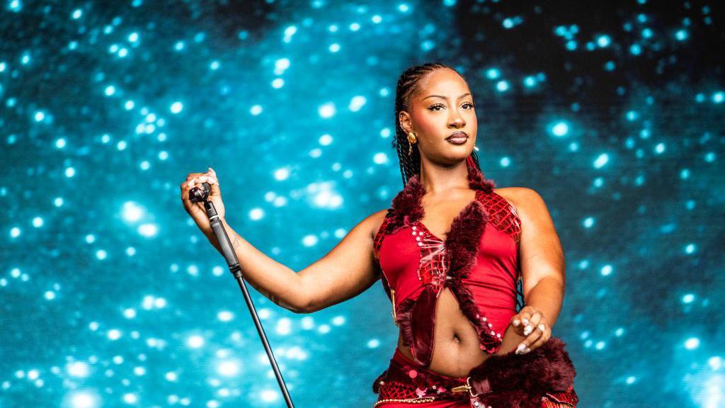 A woman holds the microphone stand and looks out tot he audience. She is wearing a red, fur-trimmed outfit. The background is a Space-like blue colour.