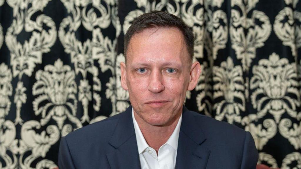 Peter Thiel wearing a white shirt and blue jacket in front of a floral wall