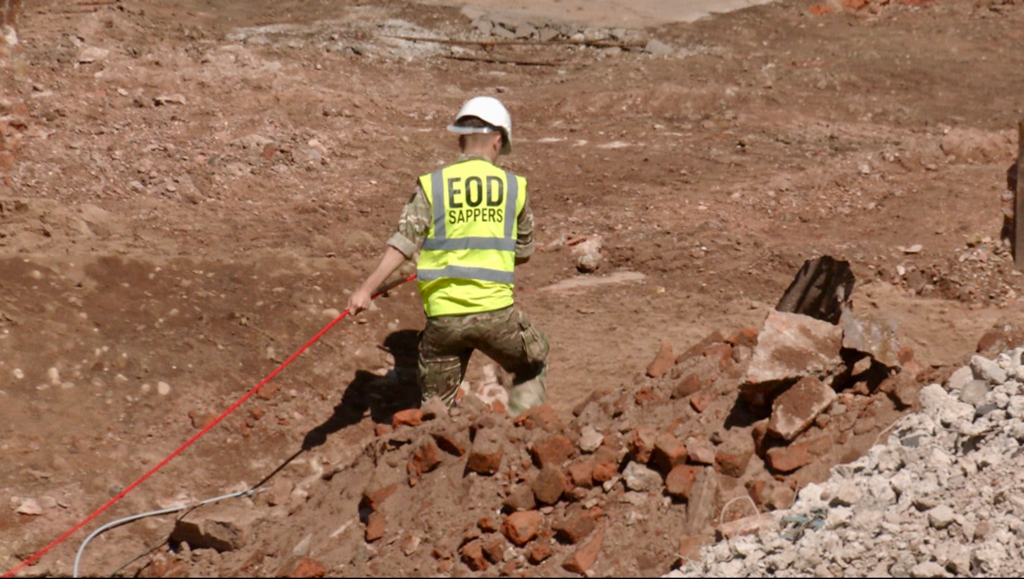 A soldier in a high-vis jacket and white hard hat pulling on a red cable on what looks like a building site