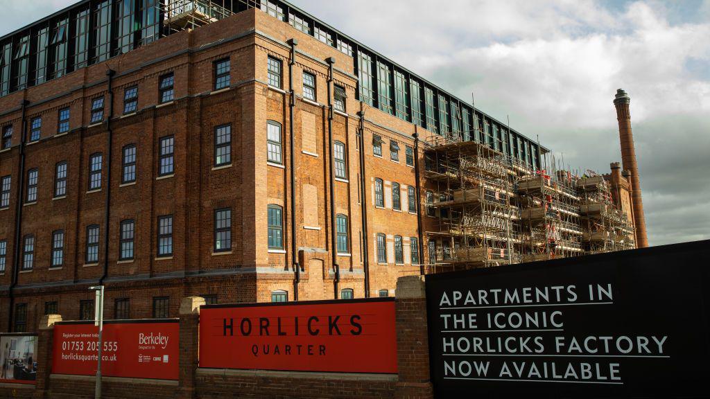 A large red-brick building, formerly a factory, being converted into flats, with scaffolding along the side. A red sign on the wall in front says 'Horlicks Quarter', and a black sign next to it reads 'Apartments in the iconic Horlicks factory now available'.