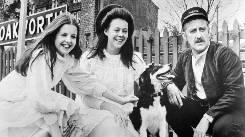 A black and white photo of actors Sally Thomsett and Jenny Agutter meeting Bernard Cribbins in a scene from the film The Railway Children. All three are crouched down on a railway station platform next to a dog.
