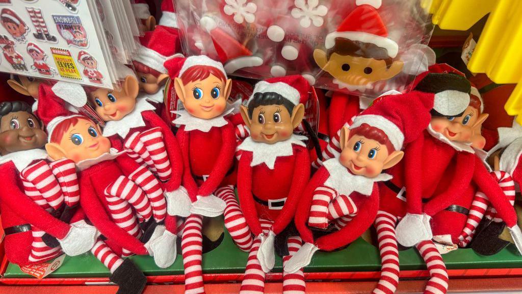 A shelf full of Elf on the Shelf toys. The elves are dressed in a red long sleeved top with red and white striped bottoms. 