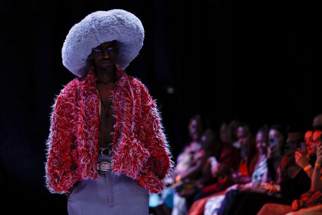 A model walks down a fashion runway wearing a huge, fluffy, lilac hat and a fuzzy red-and-white jacket. 