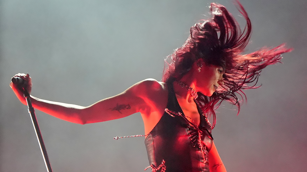 Dua Lipa on stage at Glastonbury in June. She is wearing a sleeveless top, laced up at the front. Her head is flung to the left with her hair swinging around her head in the movement. Her right arm is held out and holding on to the top of a microphone stand