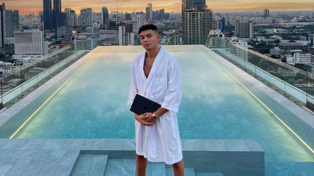 Samy Daim wearing a white dressing gown and holding what appears to be an iPad. He is standing by an infinity pool with a city skyline and sunset behind. 