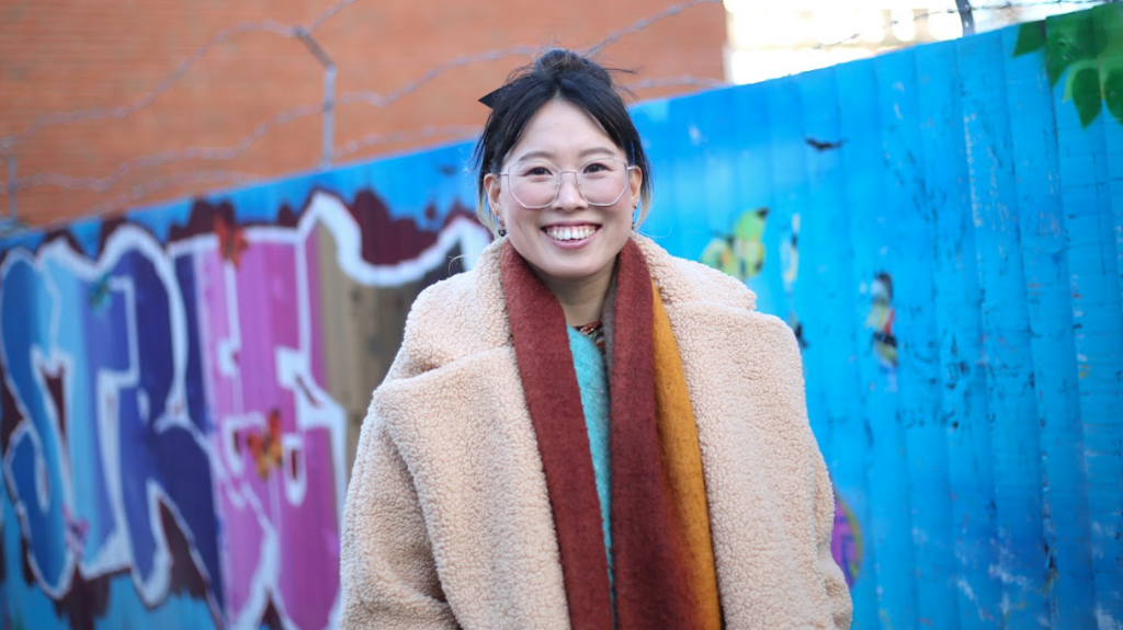 Grace wearing a thick winter coat and scarf, standing smiling in front of a fence with brightly-coloured street art