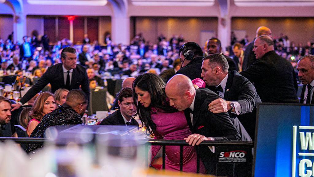 White House deputy chief of staff Stephen Miller in the foreground escorting his pregnant wife Katie away with the help of security. Wide shot of a ball room, with guests in gowns and tuxedos crouching on the ground