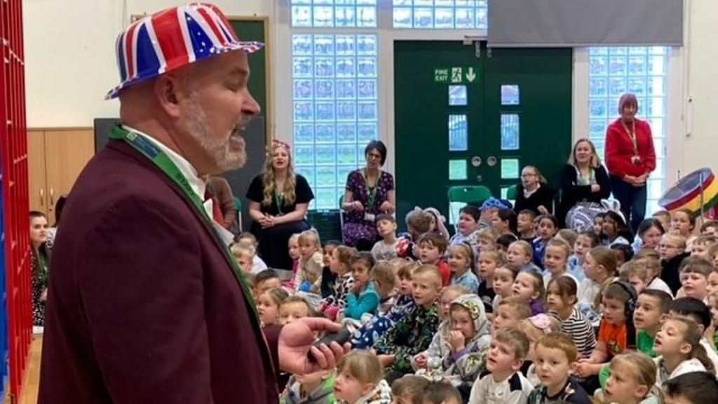A man with a purple blazer and white shirt is standing at the front of a school assembly wearing a hat decorated in Great Britain colours. Dozens of children are sitting on the floor looking at him