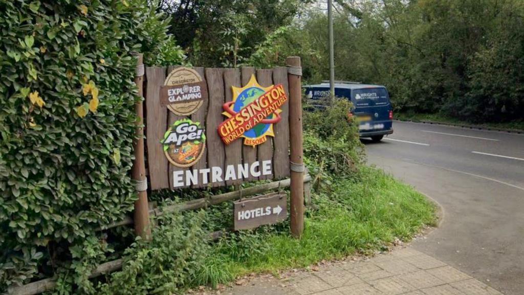 Chessington World of Adventures car park entrance with brown sign showing Chessington, Go Ape and Explorer Camping logos on them and a smaller arrow sign for the hotels.