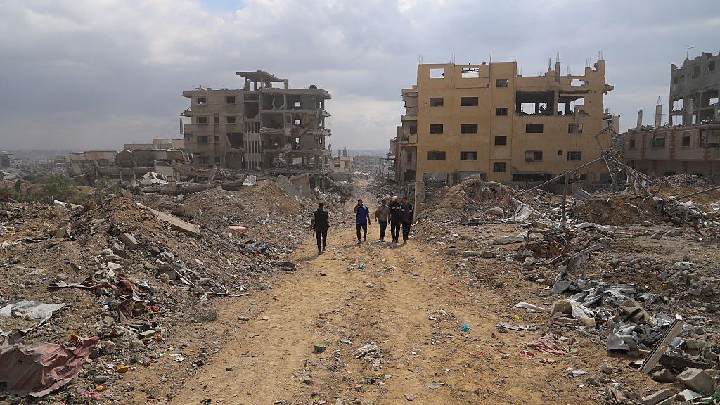 Destroyed blocks of flats and mounds of rubble line a dirt road five people are walking along in the Gabari neighbourhood of Gaza City.
