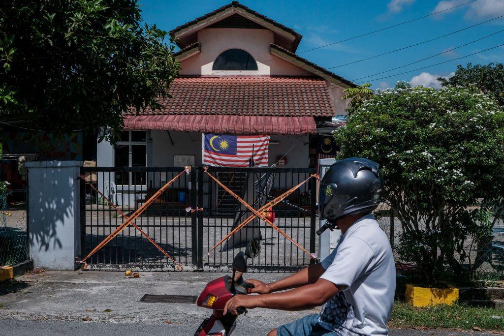 A man on a motorcyle rides past a gated building with police tape strung from the gate