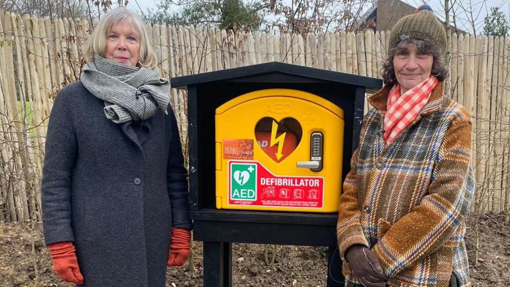 Two women wrapped up in coats, gloves and scarves are standing either side of a defibrillator, which is being kept in a wooden cabinet outside of a house.