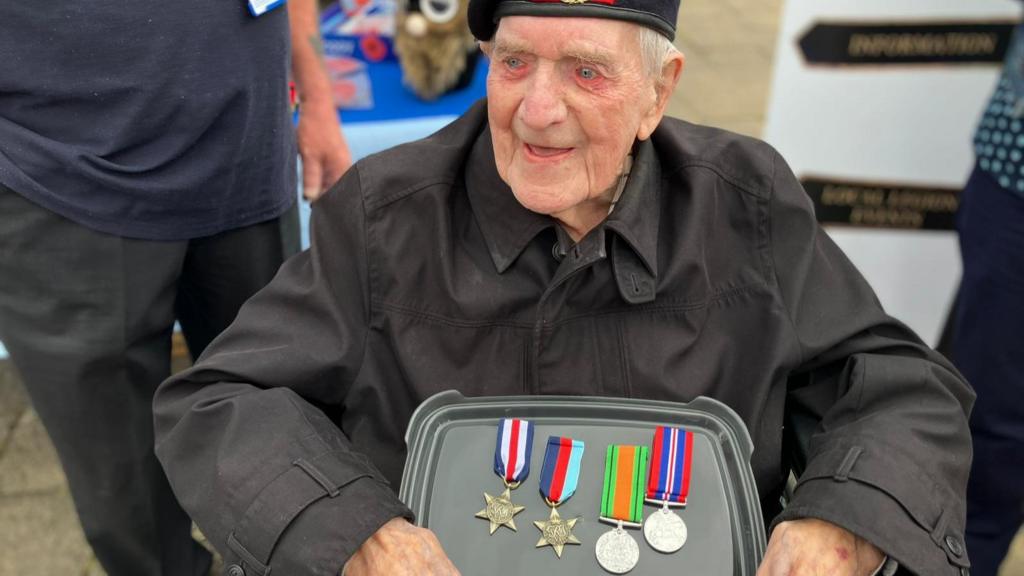 A war veteran is sat in a wheelchair holding a collection of medals. He is wearing a military beret and has tears in his eyes.